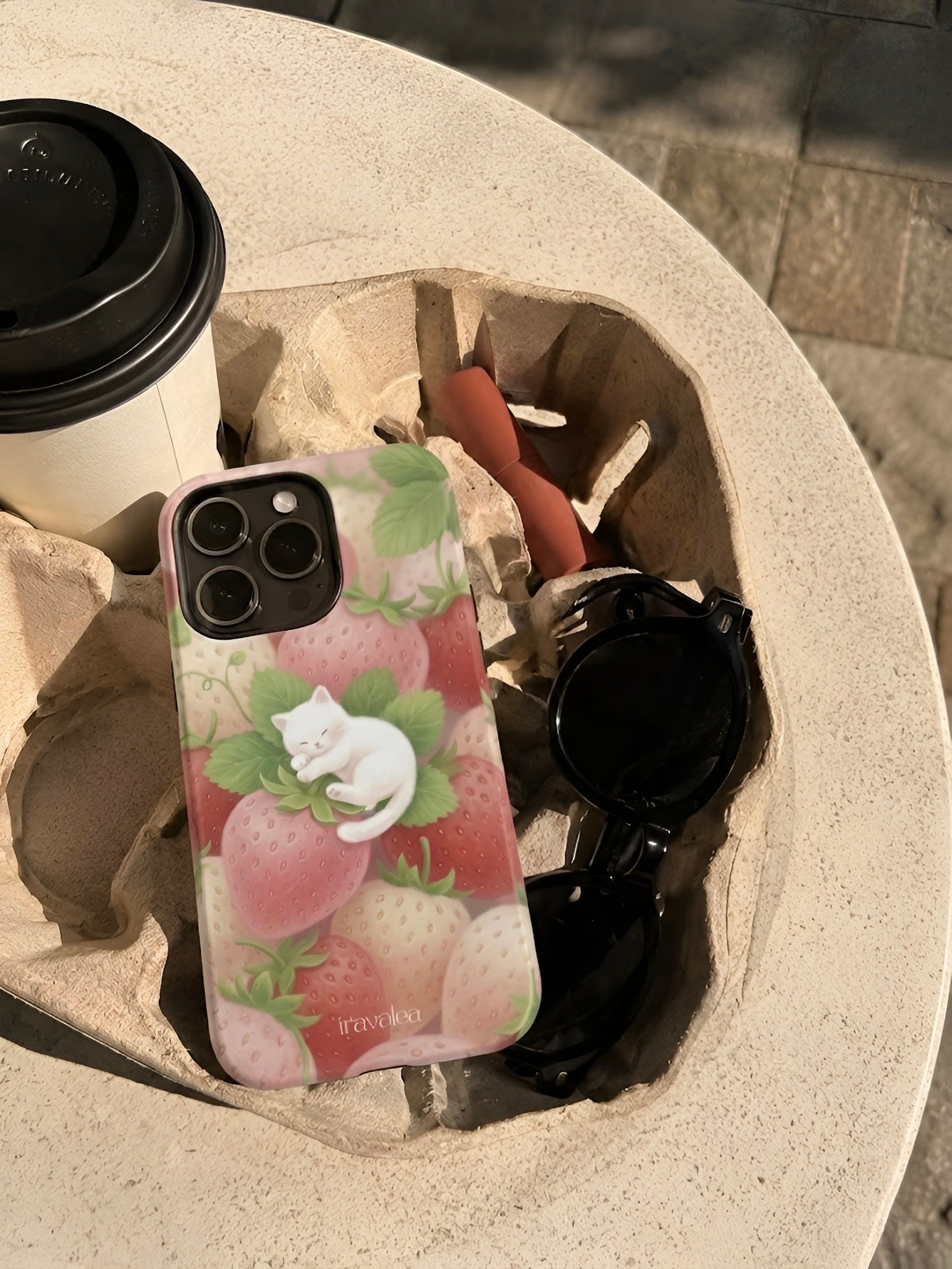Phone case with pink strawberries design on a stone surface with coffee cup and sunglasses.